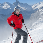 A person in a red jacket, gray pants, and a helmet with goggles stands on snowy ski slopes holding ski poles. Snow-covered mountains and a clear, blue sky form the backdrop, reminiscent of the pristine serenity one might find on a fishing trip to Tal-Adventure Lodge.