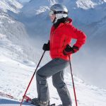 A person in a red jacket and gray pants stands on a snowy mountain slope with ski poles, wearing a helmet, goggles, and skis. Snow-covered mountains and trees are visible in the background.