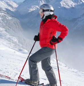 A person in a red jacket and gray pants stands on a snowy mountain slope with ski poles, wearing a helmet, goggles, and skis. Snow-covered mountains and trees are visible in the background.