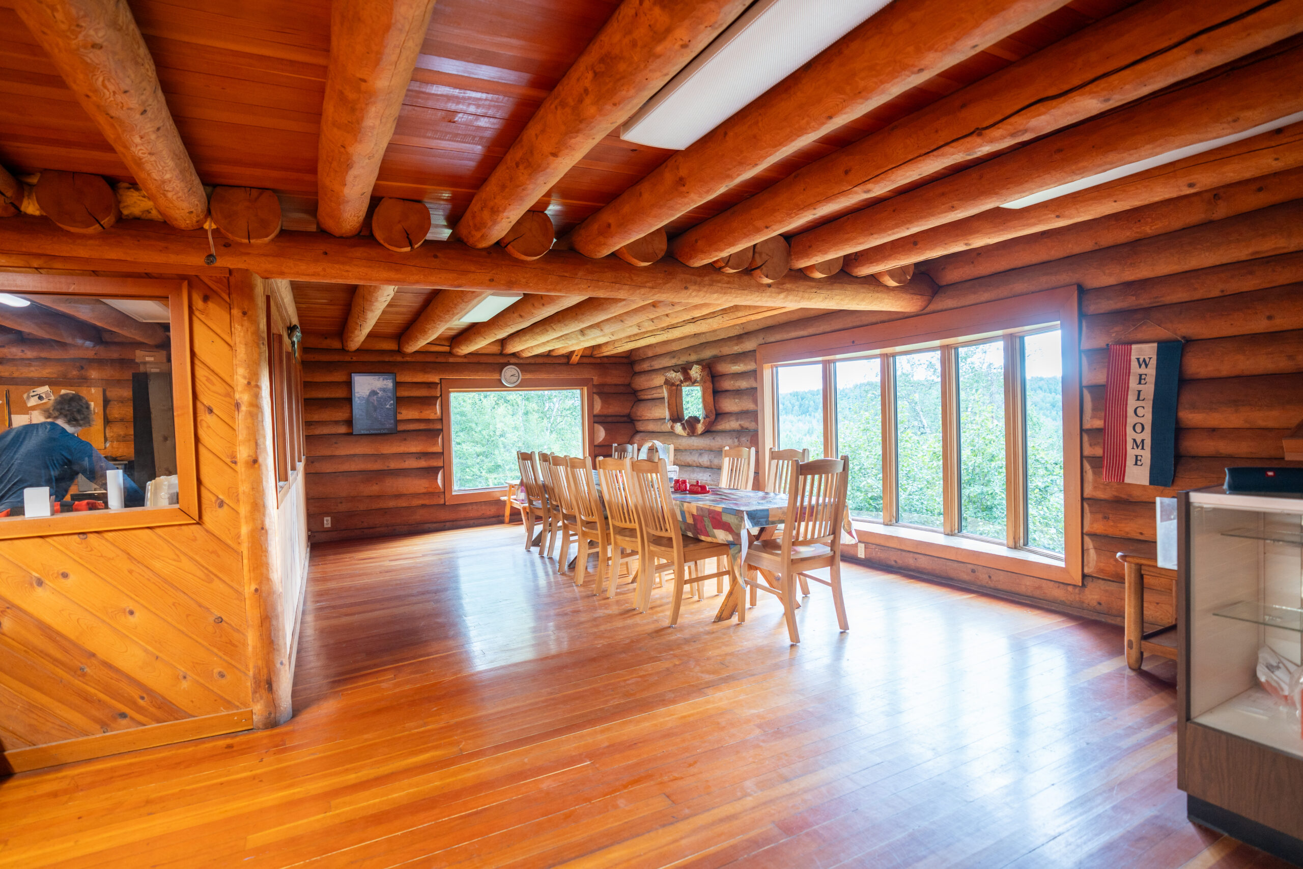 Experience a spacious dining area in the Tal-Adventures Lodge, a rustic log cabin in Alaska. Wooden walls and ceiling beams frame a large wooden dining table surrounded by chairs. Wide windows offer natural light and views of greenery, perfect after a day of fishing trips. A “Welcome” sign adorns the wall.